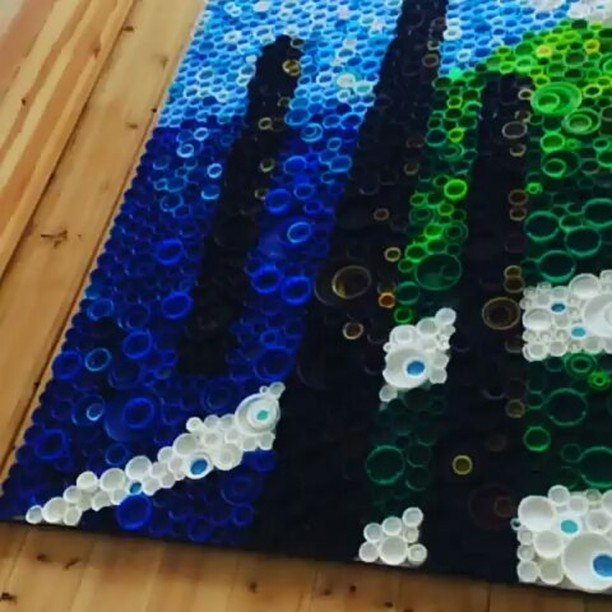 Mosaic landscape artwork made from colorful plastic bottle caps on a wooden floor.