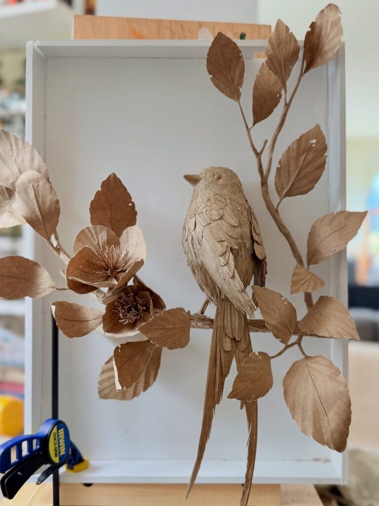 Brown paper bird sculpture perched on a paper branch inside a white frame