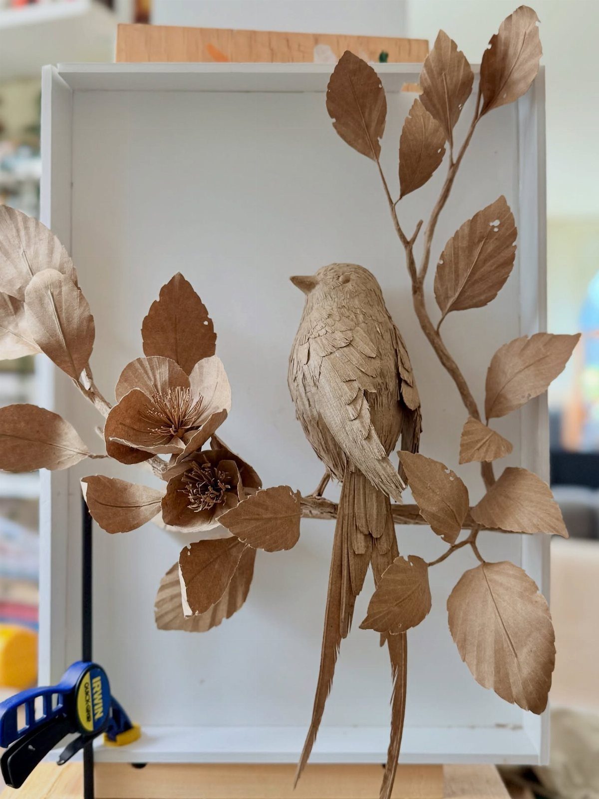 Brown paper bird sculpture perched on a paper branch inside a white frame